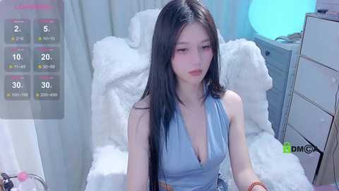 Media: Video of an East Asian woman with long black hair, wearing a low-cut blue dress, sitting on a white fur chair. Background includes a white bed and a blue-lit lamp.