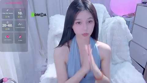 Media: A video of a young Asian woman with long black hair, wearing a light blue halter top, sitting on a plush white chair in a room with soft lighting and a large, round, pink light in the background.