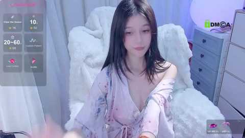 Media: Video of an East Asian woman with long black hair, wearing a floral robe, sitting on a white fur blanket in a dimly lit room.