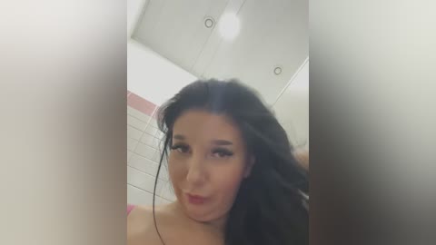 Media: A blurred video of a young woman with long, dark hair, wearing a pink top, making a pouty face in a bathroom with white tiles and recessed lighting.