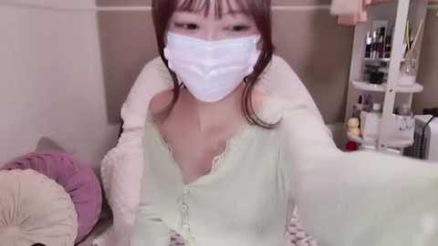 Media: Video of an Asian woman with long, straight brown hair, wearing a white face mask, light green cardigan, and a purple pillow. Background shows a messy room with a shelf full of items.