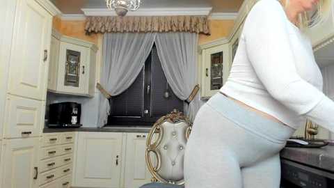 Media: A video of a woman in a white top and light gray leggings standing in a chic, cream-colored kitchen with ornate window drapes, a chandelier, and a tufted chair.