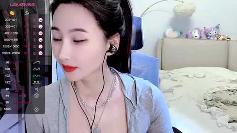 Media: A video of an East Asian woman with fair skin and long black hair, wearing red lipstick, headphones, and a gray cardigan, sitting on a bed with plush toys, in a bright room.