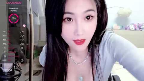 Media: Video of a young Asian woman with long black hair, wearing a light blue top, red lipstick, and a silver necklace. Background includes a heart-shaped screen, a pink flower, and a bed.