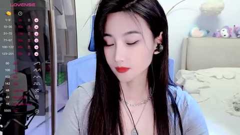 Media: A video of a young Asian woman with long, straight black hair, wearing a light grey cardigan, red lipstick, and black earrings, seated in a softly lit bedroom with a bed and shelves in the background.