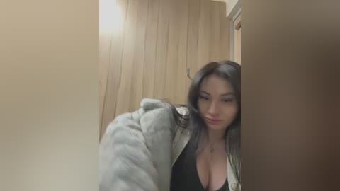 Media: Video of an East Asian woman with long black hair, wearing a grey fur jacket over a black top, posing in a dimly lit, wooden-paneled room.