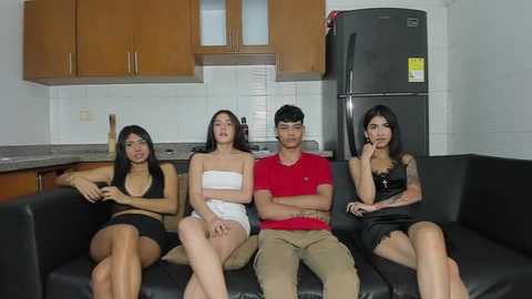 Media: Video of four young Asian adults, two women and two men, seated on a black leather couch in a modern kitchen with wooden cabinets and a refrigerator, wearing casual clothes.