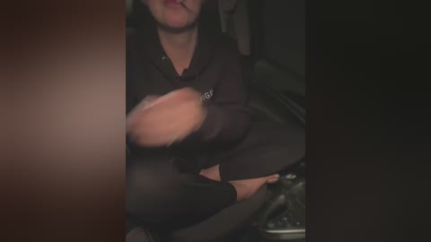 Media: A blurry video showing a person with light skin and dark hair, wearing a black hoodie, sitting on a couch. They are holding a light-colored object in their lap.