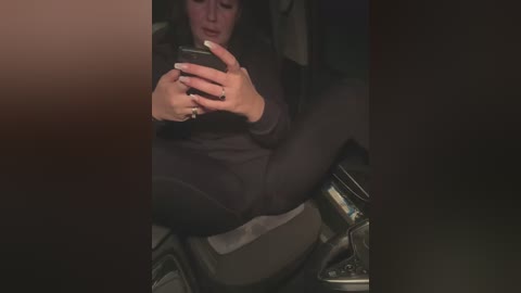 Media: A video of a woman with long black hair, wearing a black sweater, sitting in a car, engrossed in a smartphone, with a metallic car cup holder and steering wheel visible.