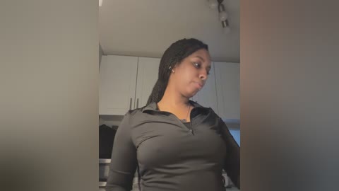 Media: Video of a Black woman with medium skin tone and braided hair, wearing a fitted, long-sleeved black top, standing in a modern kitchen with white cabinets and a stainless steel sink.