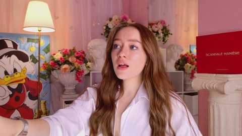 Media: Video of a fair-skinned woman with long brown hair in a white shirt, looking contemplative, in a room with pink walls, white columns, and floral arrangements.