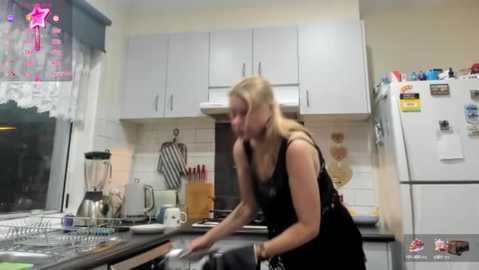 Media: A video of a blonde woman in a black dress washing dishes in a modern, well-lit kitchen with white cabinets and a stainless steel fridge.