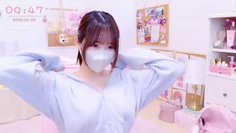 Media: A video of an Asian woman with light skin, dark hair, and a surgical mask, adjusting her blue hoodie in a pastel-themed room with pink accents and a bulletin board.