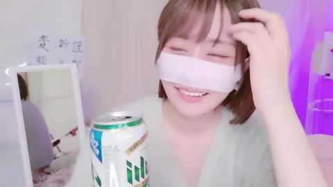 A smiling, fair-skinned, young woman with short, light brown hair wears a white face mask and a green shirt. She holds a white beer can in a dimly lit, minimalist room with a purple accent wall.