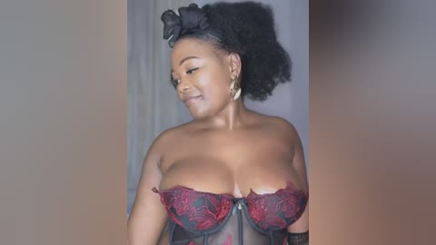 Video of a smiling Black woman with large breasts, wearing a revealing red lace corset, black hair in a bun, indoors with neutral background.