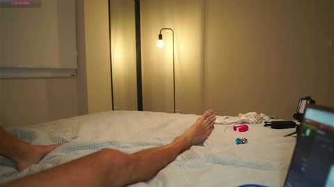 Media: Video of a person lying on a bed with white sheets, legs extended, against a beige wall with a standing lamp. Nearby, a phone and a pink vibrator are visible.