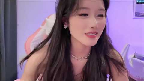 Media: Video of a smiling East Asian woman with long black hair, wearing a strapless top and silver necklace, in a softly lit room with purple walls.