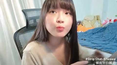 Media: Video of an East Asian woman with straight, shoulder-length brown hair and fair skin, wearing a beige shirt, sitting in a black office chair, in a messy bedroom with blue bedding and colorful toys.