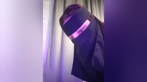 Media: Video of a person wearing a black balaclava with a slit revealing their eyes, standing in a dimly lit room with purple and pink lighting.