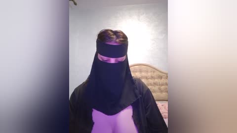 Media: Video of a woman wearing a black niqab in a dimly lit bedroom with a beige headboard and a pink bedspread.