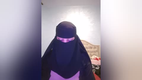 Media: Video of a woman wearing a black niqab and white shirt, standing in a dimly lit room with a white wall, a bed with red sheets, and a beige headboard.