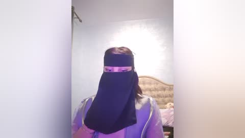 Media: Video of a person with light skin, wearing a black niqab covering the face, standing in a dimly lit bedroom with a beige headboard and soft light from an unseen source.