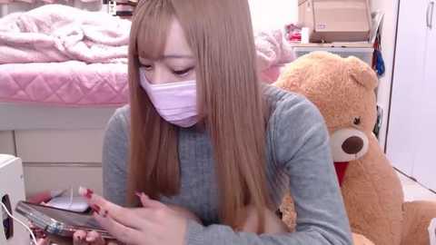 Media: Video of an Asian woman with long straight hair, wearing a face mask, seated on a bed, holding a black device, surrounded by plush toys and bedding.