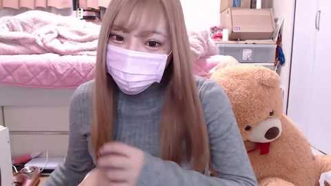 Media: Video of a young Asian woman with long straight brown hair, wearing a face mask, grey sweater, and sitting on a bed with a plush teddy bear, surrounded by cluttered bedroom items.