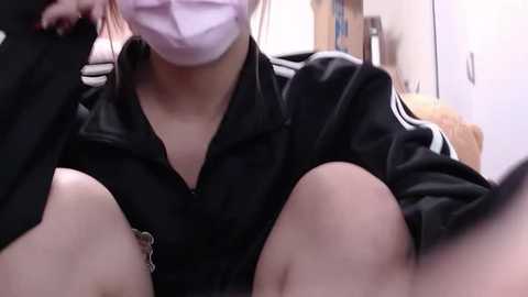 Media: Video of a person wearing a black jacket and purple surgical mask, with their legs visible and a bed in the background.