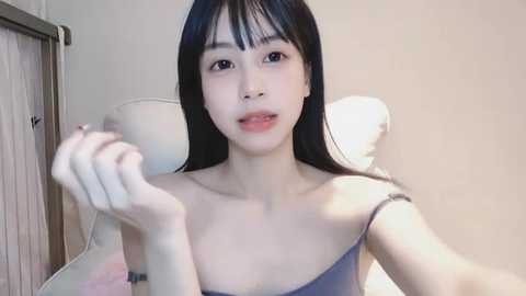 Media: A video of an East Asian woman with long black hair, fair skin, and delicate facial features, wearing a light blue spaghetti strap top, seated on a white chair in a minimalistic room.