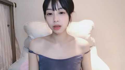 Media: Video of an East Asian woman with fair skin, black hair in a bun, wearing a sleeveless, off-shoulder purple top, sitting on a white chair in a minimalist room with beige walls and a wooden door.