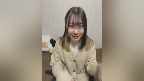 Media: A video of an Asian woman with straight, dark hair, light skin, and a slight smile, wearing a beige cardigan over a white top, kneeling on a brown carpet, surrounded by a white wall with subtle vertical lines and a stuffed animal in the background.