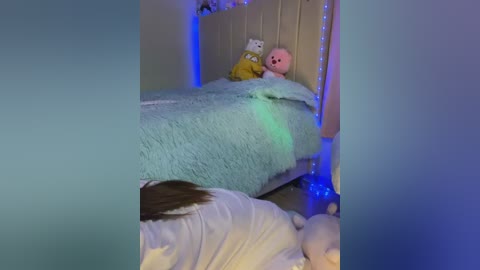 Media: Video of a dimly lit bedroom with a bed covered in a light blue, fuzzy blanket. A stuffed pink bear and a white cat toy sit on the bed. A person with long brown hair lies on the bed, wearing a white shirt.