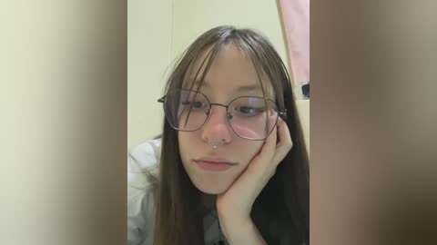 Media: A video of a young woman with long brown hair, glasses, and a septum ring, resting her head on her hand, in a bathroom with beige walls and a pink towel.