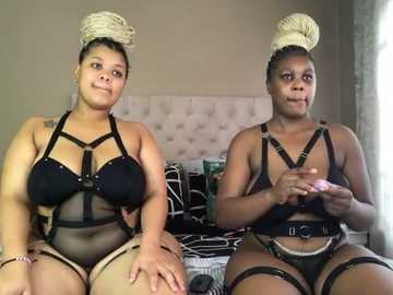 Media: Video of two plus-sized Black women in revealing black harness lingerie with matching beige hair buns, sitting on a bed with white and black bedding, in a dimly lit bedroom.