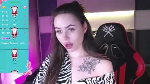 Video of a young woman with long brown hair, wearing a black and white zebra print off-shoulder top, seated in a red and black gaming chair, with a detailed black floral tattoo on her right shoulder.