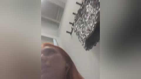 Media: Video of a blurred person with light skin and reddish hair, looking up, against a background with a chandelier and a wall with hooks.