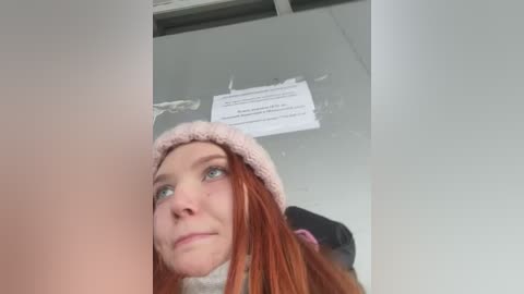 Media: Video of a young, pale-skinned woman with long red hair, wearing a pink knit hat, looking upwards, with a sign on a ceiling above her.