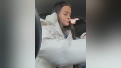 Media: A video of a young Black woman with long straight hair, wearing a white coat and seated in a car. She is texting on her phone. The background is blurred.