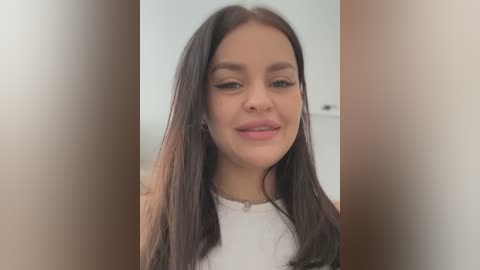 Media: A close-up video of a young woman with straight, shoulder-length dark brown hair, light skin, and a slight smile. She wears a white shirt. The background is blurred, focusing attention on her face.