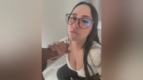 Media: Video of a fair-skinned woman with long dark hair, wearing glasses, and a low-cut white top, kneeling and sucking a man's erect penis.