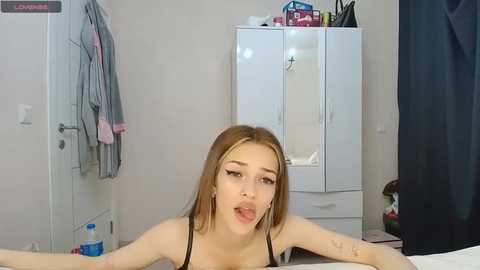 Media: A video of a young woman with fair skin, light brown hair, and medium-sized breasts, wearing a black bra, lying on a bed with a white wall, closet, and clothing in the background.