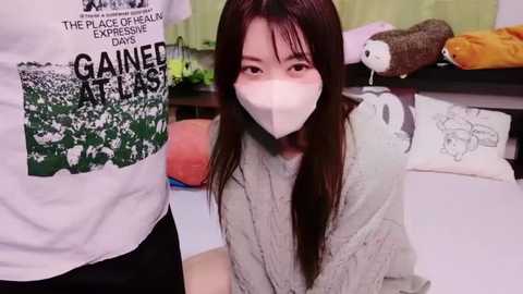 Media: Video of an East Asian woman with long black hair and a white mask, wearing a gray sweater, in a cluttered room with art supplies and a whiteboard.