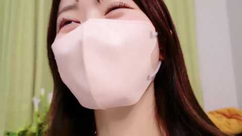 Media: Video of an Asian woman with long, straight brown hair, wearing a white surgical mask, eyes closed, smiling, with a blurred green curtain and white wall in the background.