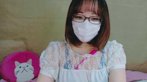 Media: Video of an East Asian woman with short brown hair, wearing glasses, a white mask, and a white lace dress with floral patterns, standing against a beige wall with a pink cat cushion.