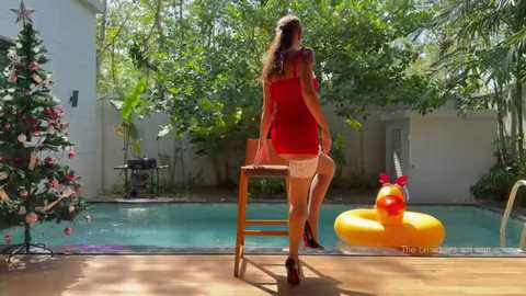 Media: Video of a woman in a red dress sitting on a stool by a pool, surrounded by lush greenery and a decorated Christmas tree.