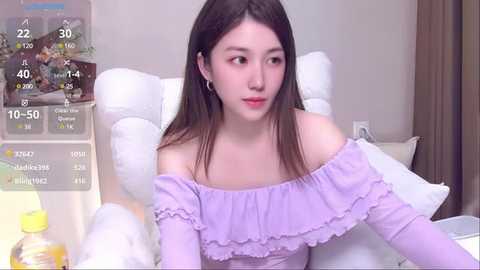 Media: Video of a young East Asian woman with straight brown hair, fair skin, and light makeup. She wears an off-shoulder lavender dress, sitting on a white chair. Background includes a weather app and a white pillow.