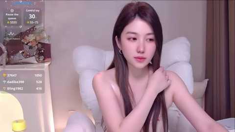 Media: Video of a young Asian woman with long dark hair, bare chested, sitting on a bed, wearing headphones, looking at a camera in a dimly lit room.