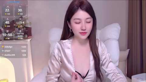 Media: A video of a young Asian woman with long, straight brown hair, wearing a satin robe, sitting on a bed, with a digital clock showing 10:50 and a temperature of 23 degrees Celsius.