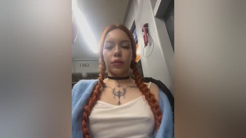 Media: Video of a young woman with long, braided, red hair, wearing a white tank top and blue cardigan, with visible tattoos on her chest. She's in a dimly lit, narrow hallway with a fluorescent light and door signs.